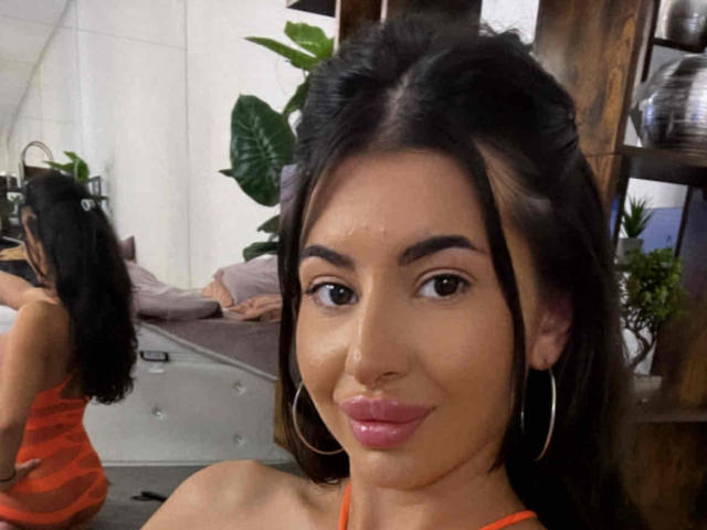 LatinaLoco with dark hair, hoop earrings, pink lipstick, and brown eyes, wearing an orange top, smiling at the camera, reflection in the mirror on the left.