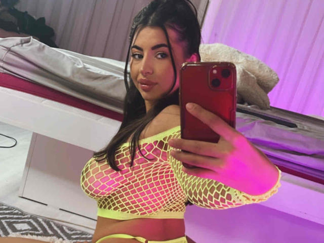 LatinaLoco with big tits wearing a neon yellow fishnet top, holding a red phone, taking a selfie, in a bedroom with pink lighting.