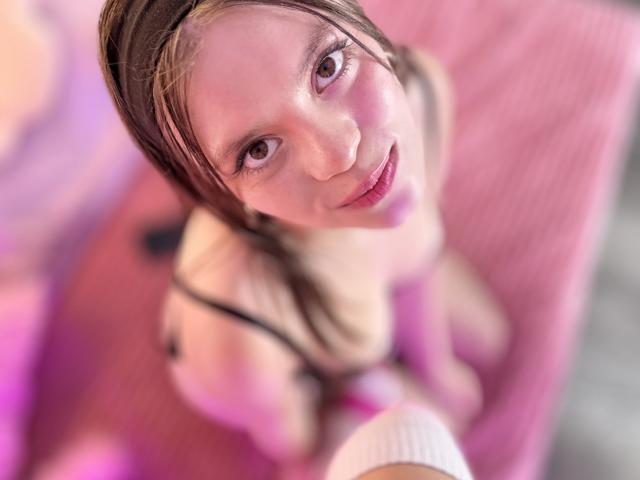AsianAndLatina with pink eyes, brown hair, and a slight smile, wearing black straps, looking up, top-down shot, pink and white background, visible white sock on camera's hand.