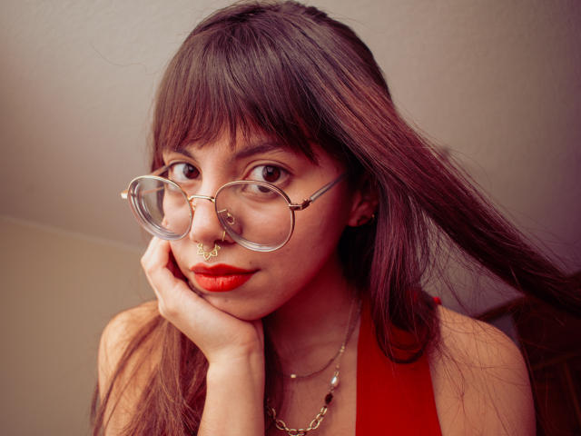 NaturalMariaHun with red lipstick, nose ring, and clear glasses, touching her cheek, wearing a red halter top, and a necklace, has dark brown hair and a cute face.