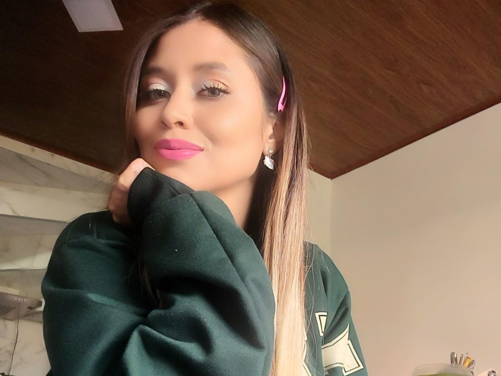 LoveCarla69 with long brown hair, pink hair clip, green sweatshirt, pink lipstick, and white earrings, smiling softly, hand near mouth, wooden ceiling above.