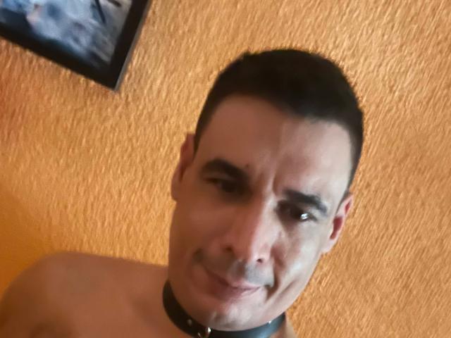 CoolManu with short black hair, wearing a black leather collar, smiling slightly, shirtless, against an orange textured wall. Slight wrinkle on forehead, dark eyebrows, slight smile.