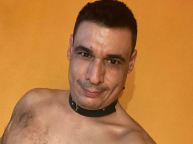 Shirtless CoolManu with a black collar, short dark hair, slight mustache, and a smile, looking at the camera, showing a hairy chest. Orange background.