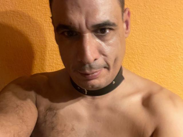 Shirtless CoolManu with a black collar, looking down, has a muscular chest, hairy skin, and a slight smirk. Orange textured wall in the background.