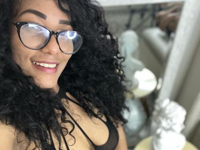 ThiainaRose, avec ses cheveux noirs bouclés, portant des lunettes, souriante, montrant ses dents blanches, un soutien-gorge noir, des seins de taille moyenne, une peau bronzée et du rouge à lèvres rose, dans un autoportrait..