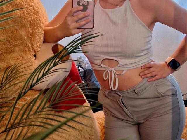 CamilaGomez with a white ribbed tank top tied at the front, showing her small tits, and gray pants, wearing a smartwatch, taking a mirror selfie.