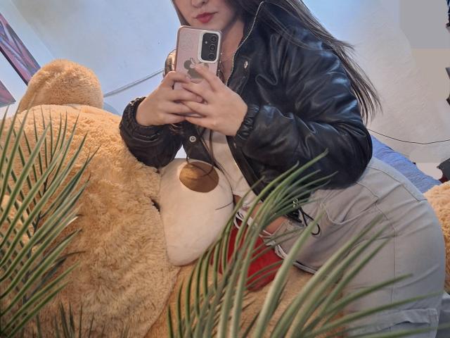 CamilaGomez with long brown hair, black leather jacket, gray pants, taking a mirror selfie on a giant tan teddy bear, holding a pink phone, red underwear visible.