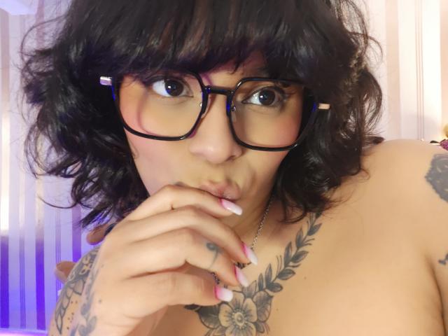 Chili com cabelo preto encaracolado e óculos, chupando o dedo, tem tatuagens no peito e no braço, e grandes mamas.