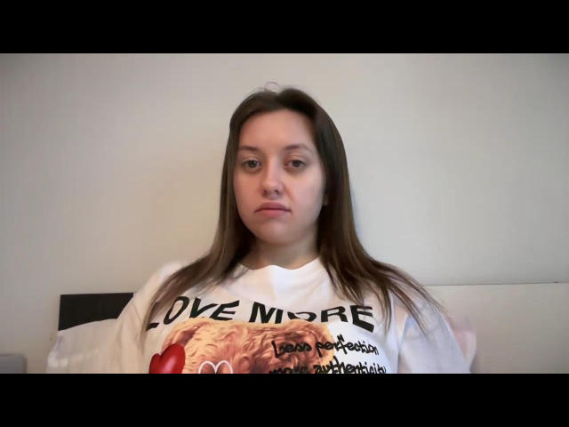 MilanaLalove with long brown hair, wearing a white shirt, sitting on a bed, looking bored, with a neutral expression, and slightly puffy ass.