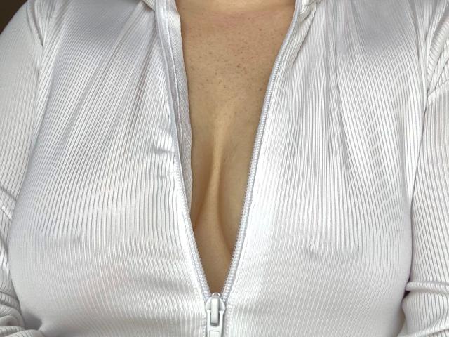 A camiseta branca com costelas e zíper da kinky69couple-hot, parcialmente aberta, revelando um decote profundo com seios grandes, redondos e levemente texturizados visíveis..