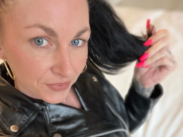 13-Mel-hot with blue eyes, holding black hair, wearing a black leather jacket, gold earrings, and pink nails, smiling slightly.