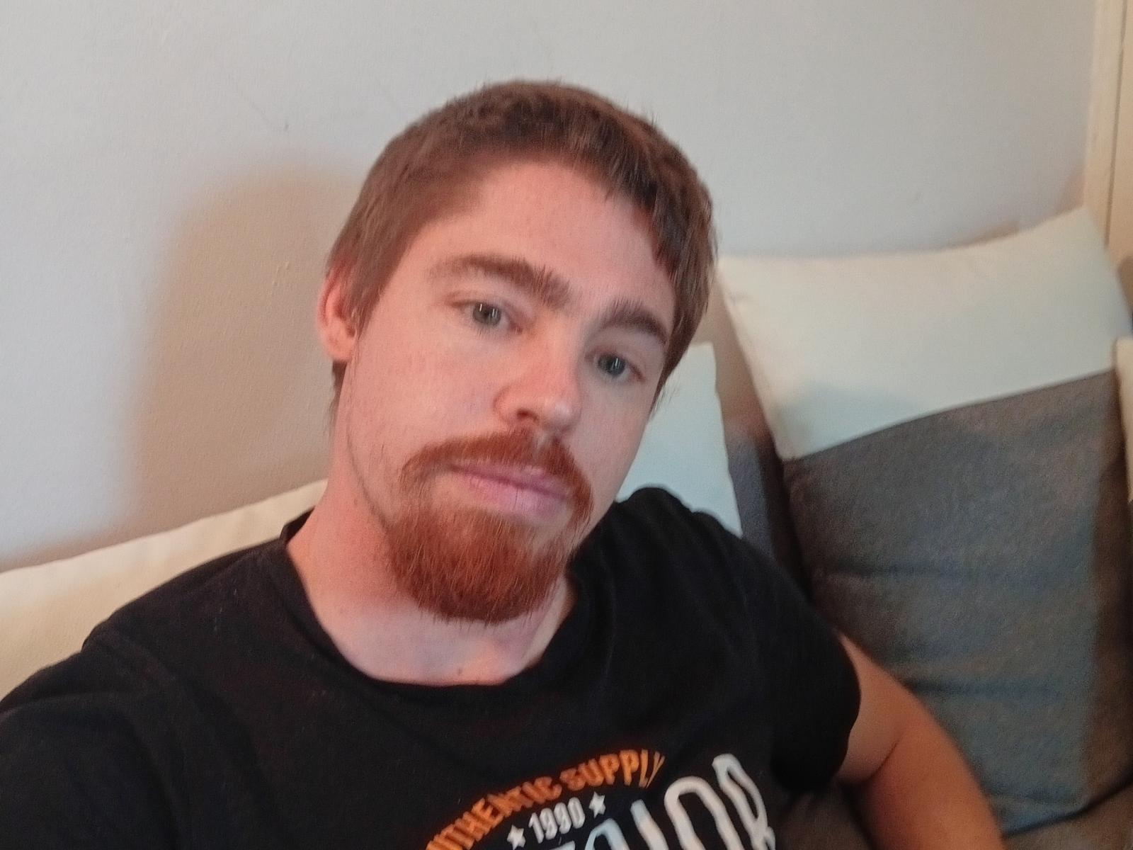 SoumiDu69 has a medium build, short brown hair, and a red beard, wearing a black t-shirt, sitting on a gray and white pillow, with a neutral expression.