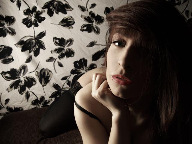 BettyBliss, con i capelli castano scuro, le labbra piene e una maglia di pizzo nero, appoggia il mento sulla mano, mostrando il seno, in una foto scura e malinconica..