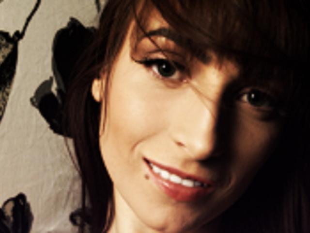 BettyBliss, con i capelli castani, gli occhi verdi e un leggero sorriso, mostra una piccola lentiggine sul labbro inferiore. Il suo viso è leggermente ombreggiato, il che ne accentua i lineamenti..