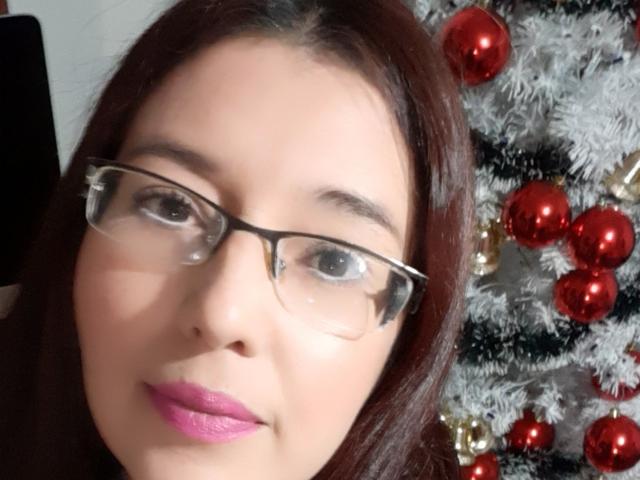 AntonnellaS has brown hair, glasses, pink lipstick, and a cute face, with a white Christmas tree adorned with red and gold ornaments in the background.