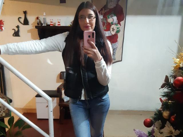 AntonnellaS with long brown hair, glasses, white shirt, black vest, blue jeans, taking a mirror selfie with pink phone, standing next to a Christmas tree, showing her ass.