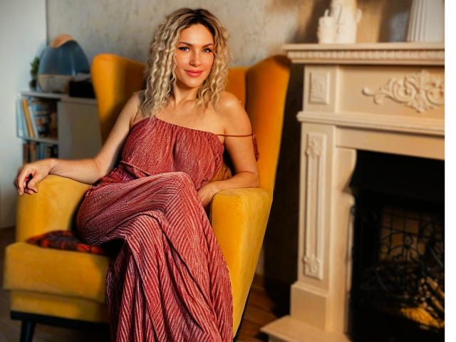 DAkotaHotti69 with curly blonde hair, off-shoulder red dress, sitting in yellow chair, hands on armrests, smiling, next to ornate white fireplace, wooden floor, cozy setting.