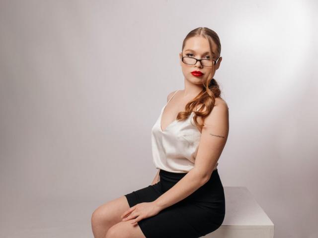 LaraPika with red lipstick, glasses, white satin top, black skirt, sitting on white cube, ponytail, tattoo on right arm, hands on knees, looking at camera, smiling.