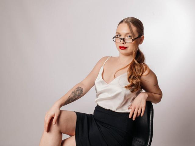 LaraPika with red lipstick, glasses, white tank top, black skirt, and tattoo on left arm, sitting on chair, legs crossed, right arm on knee, looking at camera.