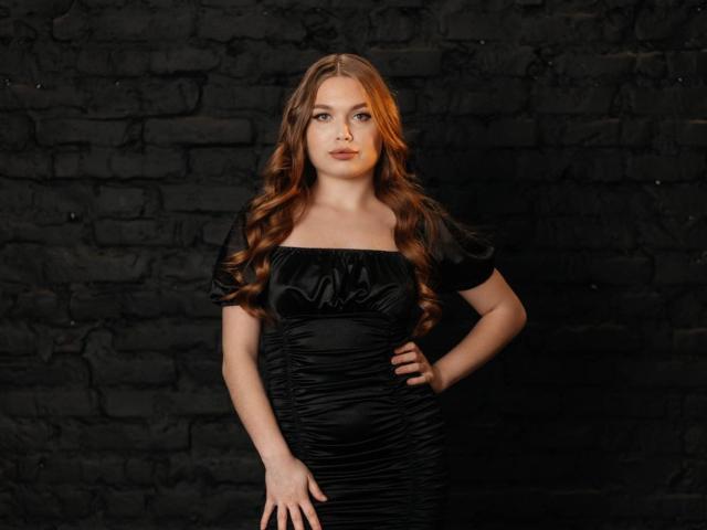 LaraPika with long red hair, wearing a black ruched dress, standing against a dark brick wall, hand on hip, looking serious, soft lighting, slight smile.