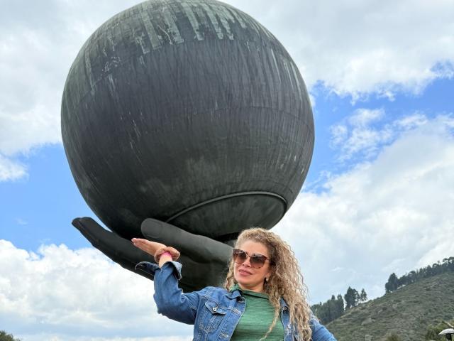Dalila, con el pelo rubio rizado, vistiendo una chaqueta vaquera y una blusa verde, está de pie frente a una esfera negra gigante, sosteniéndola con la mano levantada..