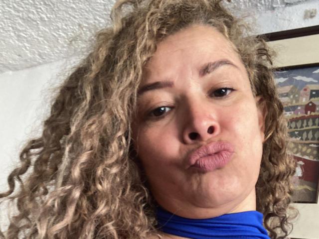 Le visage de DalilaSuperHot, avec ses cheveux blonds bouclés, ses yeux marrons et ses lèvres pincées, portant un collier bleu, mettant en valeur son nez et ses lèvres légèrement entrouvertes. Elle regarde directement l'appareil photo..