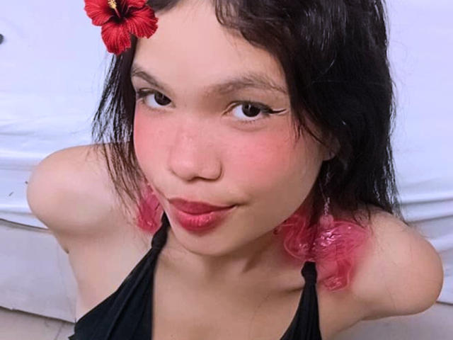MidnightHarpy with pink puffy hair, red hibiscus in hair, black halter top, and red lipstick, showing her small tits and cute face with a slight smile.
