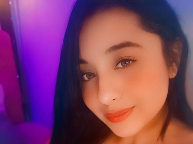 IsabelSaenzX has a cute face with light brown skin, dark eyebrows, brown eyes, and orange lipstick. She has long black hair and a subtle smile.