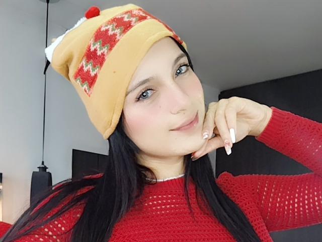 ValeryeCook with long black hair, blue eyes, and a cute smile, wearing a red knit sweater and yellow knit hat with a red pom-pom, resting her chin on her hand.