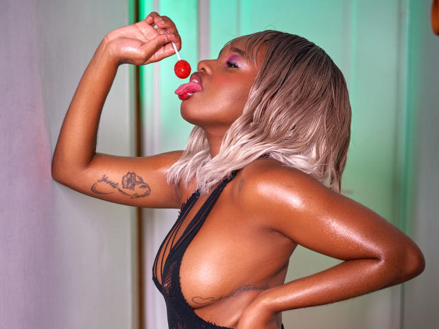 EmmaParisi with shiny brown skin, blonde bob, black lace top, tongue out, licking red candy, tattoo on arm, hand on hip, showing big tits, sexy, seductive.