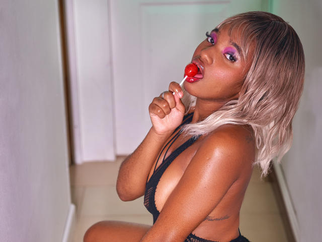 EmmaParisi with dark brown skin, blonde hair, pink eye makeup, red lipstick, sucking a red lollipop, wearing a black strappy top, showing her tits, in a hallway.