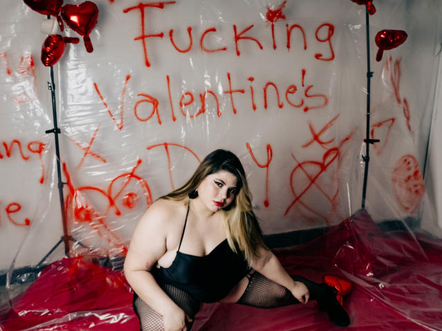 EymiTomsom with pale skin, long blonde hair, black lingerie, fishnet stockings, red lipstick, sitting on red plastic, surrounded by red heart balloons, looking at camera.