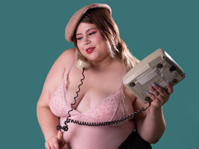 EymiTomsom, chubby with pale skin, wearing pink lace bra, beige beret, holding vintage phone with coiled cord, smiling, pink lipstick, teal background, visible large tits, holding phone.