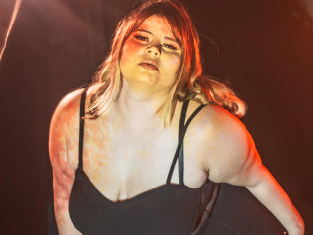 EymiTomsom with large boobs in a black dress, looking annoyed, has red and orange lighting on her, and her blonde hair is slightly messy. Her skin is pale.