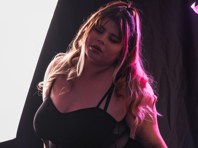 EymiTomsom with large boobs, wearing a black top, has long wavy blonde hair with pink highlights, eyes closed, and a slightly open mouth, standing in front of a light source.