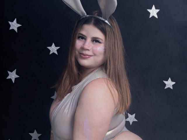 EymiTomsom with brown hair, wearing white bunny ears and a shiny, tight, sleeveless top, showing her ass, smiling at the camera, face marked with white glitter.