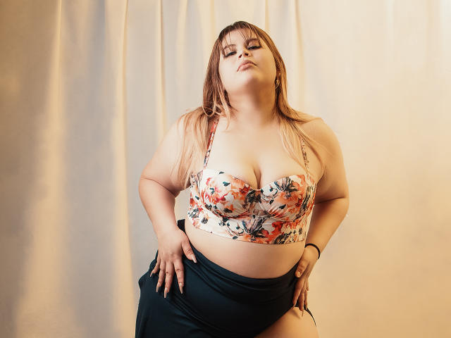 EymiTomsom, avec de grandes seins, portant un soutien-gorge fleuri, une jupe noire et de longs cheveux blonds, se tient debout avec assurance, les mains sur les hanches, et regarde vers le bas avec un léger sourire..
