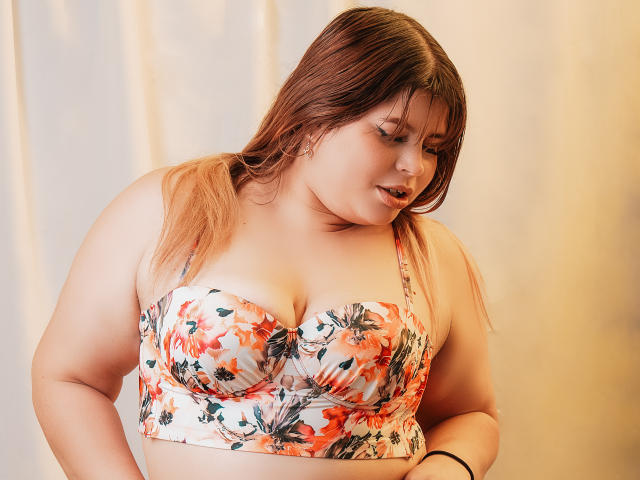EymiTomsom has big, fluffy tits in a floral bra, looking down with a soft smile. She's got light brown hair and a cute nose piercing. Her arms are relaxed.