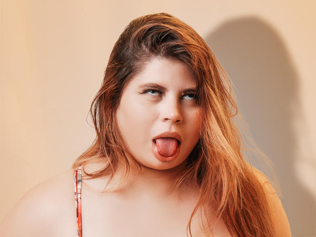EymiTomsom a de longs cheveux blonds, presque châtains, des yeux bleus, et elle tire sa langue rose. Elle porte un haut rouge avec de fines bretelles, et il y a une ombre sur la droite..