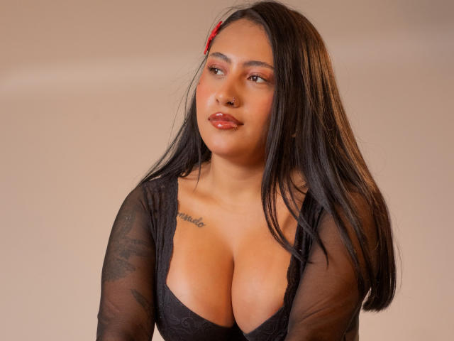 MadyBrown, con cabello largo y negro, un sujetador rojo, una blusa de encaje negro, busto grande, piel morena, un tatuaje sobre el pecho izquierdo, labios rosados brillantes, mirando ligeramente hacia la derecha..