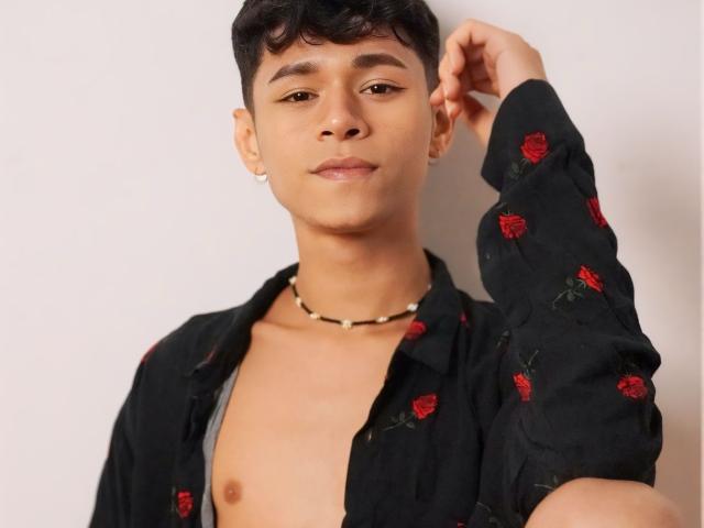 DanteEvans, shirtless, black floral shirt, small red roses, brown skin, short curly black hair, subtle makeup, gold earring, necklace, right arm raised, confident expression.
