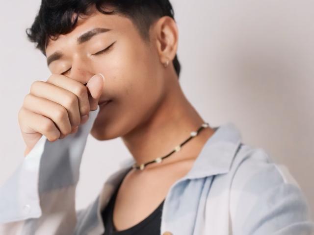 DanteEvans, with eyes closed, holds a white cloth to his nose, wearing a black shirt, white plaid shirt, and a bead necklace, looking slightly distressed.