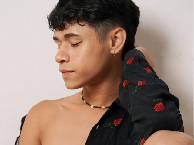 DanteEvans with short, wavy black hair, wearing a black shirt with red rose embroidery, and a bead necklace, eyes closed, left arm raised, small gold earring in right ear.