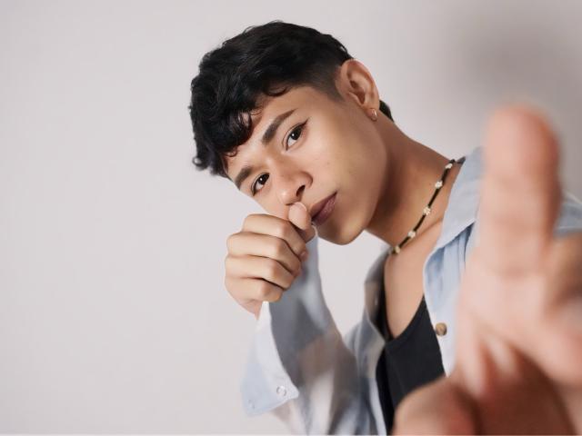 DanteEvans with short black hair, wearing a light blue shirt, black tee, and bead necklace, pointing at camera, finger on chin, serious expression, soft light, short curly hair.
