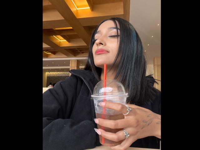SalomeMille with black hair, pink lipstick, and a confident smile, holding a cup with a red straw, wearing a black jacket, showing tattoos on her hand.