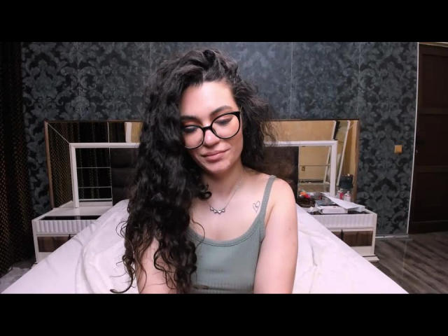 CurllyMya con capelli neri ricci, occhiali, canotta verde, seno piccolo, tatuaggio sul petto, sorridente, seduta sul letto, coperta bianca, carta da parati floreale scura, comodino con oggetti..