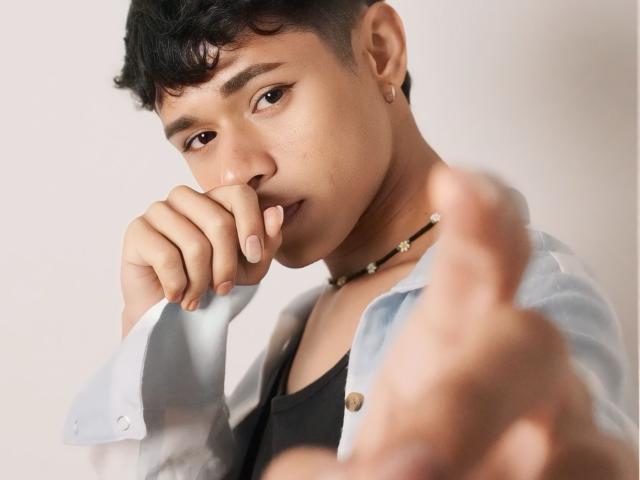 DanteEvans with short black hair, light skin, and a small earring, biting his finger, wearing a light blue shirt and black tank, holding a blurred finger in front.