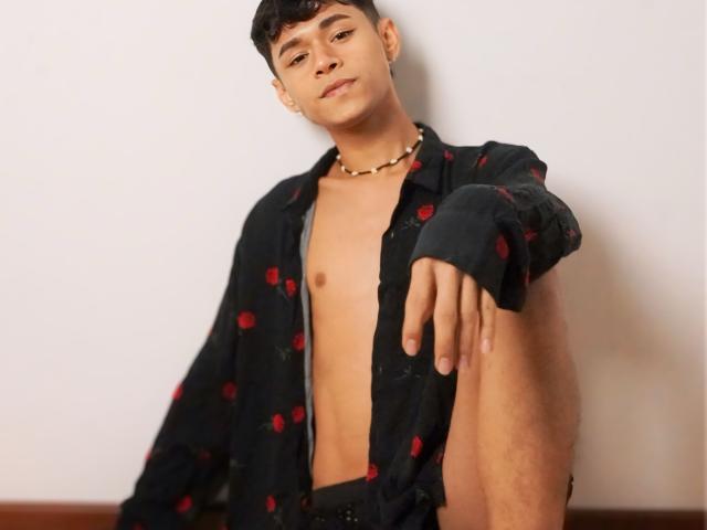 DanteEvans, shirtless, wearing black polka-dot shirt, red dots, necklace, short dark hair, slight smile, arm extended, knee up, bare chest, casual, confident, sitting, looking at camera.