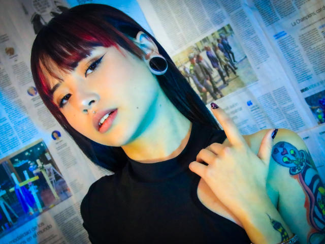 EmmahGreys with black and red hair, blue skin tone, large black ear gauges, black sleeveless top, tattooed arm, finger pointing to neck, blue and white lighting.