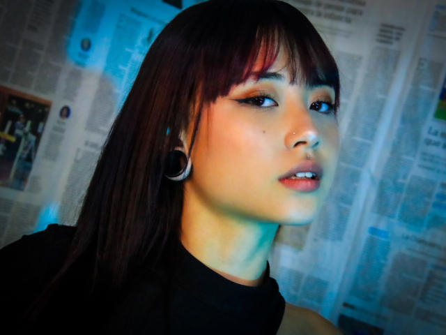 EmmahGreys has dark brown hair, black ear gauges, and is wearing a black turtleneck. She's looking to the side with subtle makeup and a small nose piercing.